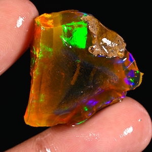 May include: A vibrant, raw opal gemstone with a mix of colors. The opal displays a striking play-of-color, with flashes of green, orange, and blue. The rough stone is held between fingers, showcasing its natural, unpolished state.