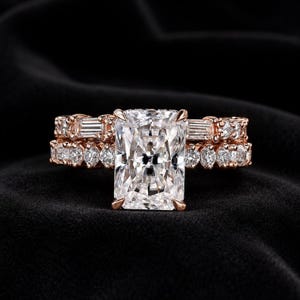 May include: A rose gold engagement ring set featuring a large rectangular radiant-cut center stone. The band is adorned with a row of baguette-cut diamonds and smaller round brilliant-cut diamonds, creating a layered, sparkling appearance.