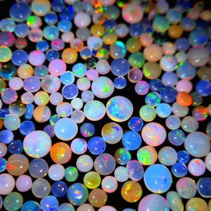 May include: A collection of opals in various sizes and colors, including blue, orange, yellow, and white. The opals have a glossy finish and display iridescent flashes of color. The background is black, highlighting the gemstones.