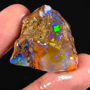 May include: A rough opal gemstone with a brown matrix. The opal displays vibrant colors, including blue, green, orange, and purple. The gemstone is held between fingers, showcasing its natural, unpolished state. The opal's surface has a glossy appearance.