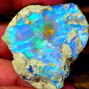 Peut inclure: Une pierre d'opale brute présentant un éventail de couleurs vives. L'opale affiche des bleus, des verts et des jaunes, avec une structure cristalline. La pierre est sertie dans une matrice de roche beige, mettant en valeur sa forme naturelle.
