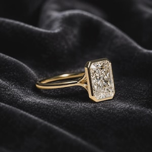 May include: A gold ring featuring a large, rectangular radiant-cut center stone held in a sleek bezel setting. The band is thin and polished, connecting to the setting with a split-shank design that highlights the clear, faceted gemstone.