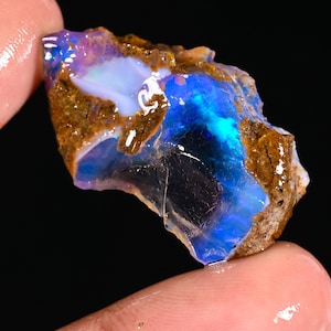 May include: A rough, uncut opal gemstone with vibrant blue, purple, and white internal colors. The stone has a dark brown matrix and is held between two fingers, showcasing its natural, raw beauty. The opal's surface reflects light, creating a shimmering effect.