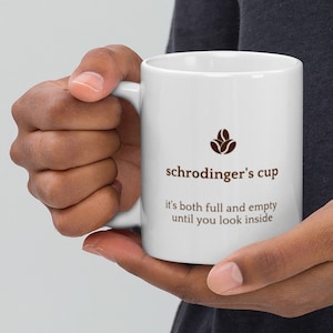 Può includere: Una tazza da caffè in ceramica bianca con un design a chicco di caffè marrone e il testo "schrödinger's cup it's both full and empty until you look inside".