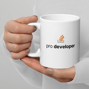 Könnte beinhalten: Weiße Keramiktasse mit einem schwarzen und orangefarbenen Logo, auf dem "pro developer" steht. Das Logo ist eine stilisierte Darstellung eines Bücherstapels.