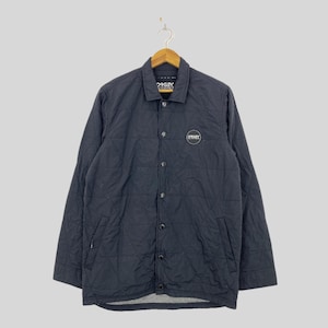 May include: A black quilted jacket with a button-up front and long sleeves. The jacket features the "Oakley" logo on the chest and collar. The jacket is hanging on a wooden hanger.