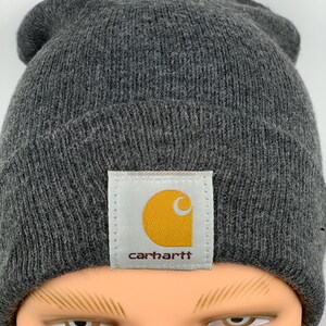 Puede incluir: Un gorro de punto gris con un parche de logo Carhartt amarillo y marr&oacute;n.