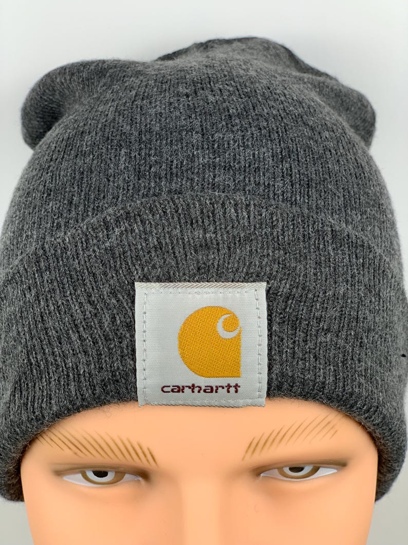 Puede incluir: Un gorro de punto gris con un parche de logo Carhartt amarillo y marr&oacute;n.
