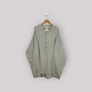 Puede incluir: Una camisa verde claro de manga larga con cuello mandarín y tapeta abotonada. La camisa tiene un bolsillo en el pecho y botones de madera. La camisa está colgada de una percha de madera.