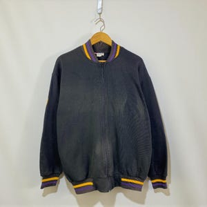 May include: Black zip-up jacket with a ribbed collar, cuffs, and hem. The collar, cuffs, and hem feature a purple and gold striped design. The jacket is hanging on a wooden hanger against a plain white background.