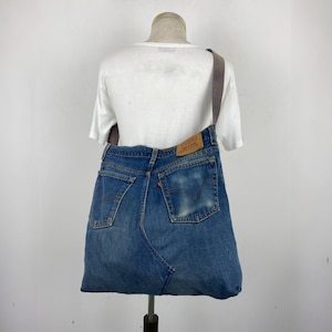 May include: A denim tote bag made from repurposed blue jeans. The bag features the back pockets and waistband of the jeans, with a brown strap and a Levi's brand patch. The bag is displayed on a mannequin.