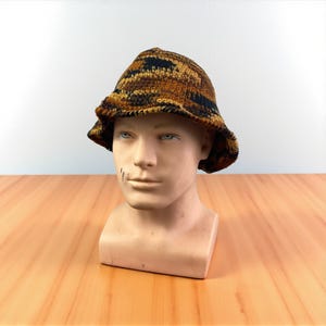 May include: A brown, black, and tan crocheted bucket hat. The hat is displayed on a mannequin head. The hat has a wide brim and a textured appearance. The hat is made of a yarn with a camouflage-like pattern.
