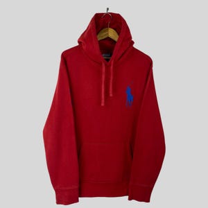 Puede incluir: Una sudadera con capucha roja con bolsillo de canguro y capucha con cordón. La sudadera presenta un logotipo de jugador de polo bordado en azul en el pecho. La prenda está colgada en una percha de madera sobre un fondo gris claro.