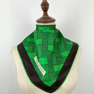 Pode incluir: Um lenço quadrado verde com um padrão geométrico preto e uma borda marrom escura. O lenço está drapeado sobre um manequim e apresenta o texto "PIERRE BALMAIN" em uma etiqueta branca.