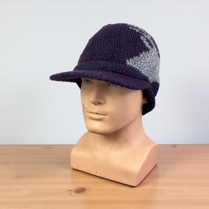 May include: A navy blue knitted cap with a small peak. The cap has a geometric design in grey and red accents. The hat is displayed on a mannequin head against a neutral background.