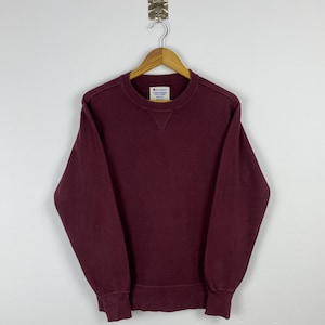 Puede incluir: Una sudadera Champion de color granate cuelga de una percha de madera contra una pared blanca. La sudadera de cuello redondo tiene un diseño sencillo con puños y dobladillo acanalados. La etiqueta con el logotipo de Champion es visible en el cuello.