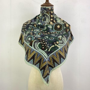 Puede incluir: Un pañuelo cuadrado con un patrón complejo en tonos verdes, azules y marrones. El pañuelo presenta un borde geométrico con un diseño en zigzag en marrón y dorado. El texto "Guy Laroche Paris" es visible en el pañuelo.