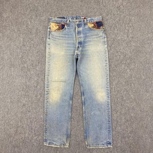 May include: Faded light blue denim jeans with floral patches on the front pockets. The jeans feature a button fly and a straight-leg cut. The jeans have a worn, vintage look.