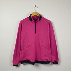 May include: A magenta-colored long-sleeved pullover with a half-zip closure and a black collar and hem. The pullover has two front pockets and a small logo on the lower right side. The garment is hanging on a wooden hanger.