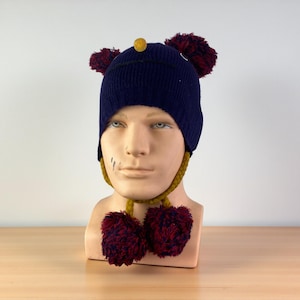 May include: A navy blue knit hat with two burgundy pom-poms on top and two more at the ends of the mustard-colored ties. A small yellow pom-pom is centered on the hat. The hat is displayed on a mannequin head.