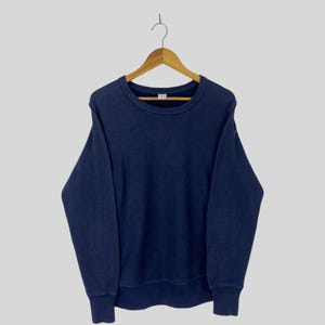 May include: A navy blue crew neck sweatshirt hanging on a wooden hanger. The sweatshirt has long sleeves and ribbed cuffs and hem. The fabric appears to be a soft, textured material.