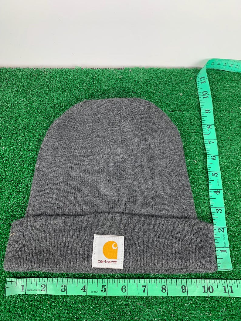 Puede incluir: Gorro de lana gris con un parche del logotipo de Carhartt. El logotipo es amarillo con un contorno negro y la palabra "carhartt" en blanco.