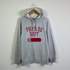 Può includere: Una felpa con cappuccio grigia con interno bianco e tasca frontale. La parte anteriore della felpa presenta la scritta "PHYS ED DEPT" in lettere rosse a blocchi sopra un rettangolo rosso con una "G" bianca all'interno.
