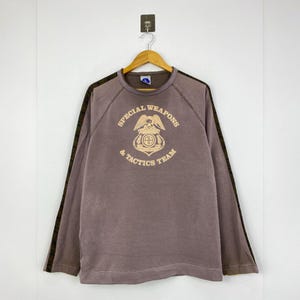 May include: A brown long-sleeved sweatshirt with the text "SPECIAL WEAPONS & TACTICS TEAM" in a circular design. The design includes an eagle and a shield. The sleeves have a dark brown stripe down the side.