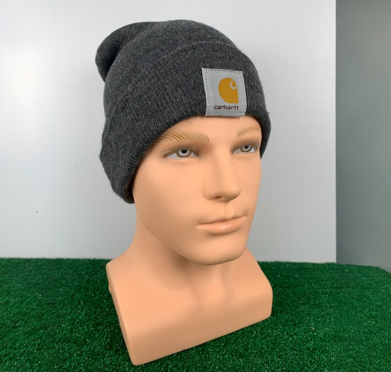 Puede incluir: Gorro gris con un parche de logotipo Carhartt amarillo. El gorro est&aacute; hecho de un tejido suave de punto.