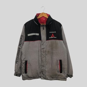 May include: A black, red, and white Mitsubishi Motors jacket with a houndstooth pattern. The jacket features the text "MITSUBISHI MOTORS" on the left sleeve and "Drive@earth" with the Mitsubishi logo on the chest. It has a red interior lining.