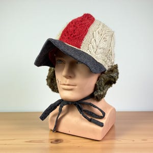 Könnte beinhalten: Ein Winterhut mit Strickmuster in Creme und Rot, mit einem dunkelgrauen Schirm und Kunstfell-Ohrenklappen. Der Hut hat einen Kordelverschluss und wird auf einem Schaufensterkopf präsentiert.