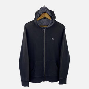 May include: Black zip-up hoodie with a hood and a small logo on the chest. The interior of the hood features a patterned lining. The hoodie has a front pocket and ribbed cuffs.