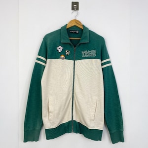 May include: A green and cream zip-up jacket with white stripes on the sleeves. The jacket has a collared neckline and features patches and the text "T&CSD" across the chest. The jacket is hanging on a wooden hanger.
