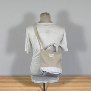 Puede incluir: Un bolso tote beige y gris con una correa larga para el hombro. El bolso tiene forma rectangular con una sección inferior blanca y gris. Una pequeña etiqueta es visible en la parte delantera del bolso. El bolso se muestra en un maniquí.