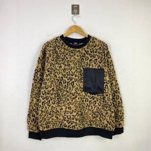 Pode incluir: Um moletom com estampa de leopardo bege e preto, com um bolso preto no peito. Possui gola, punhos e bainha pretos. A peça está pendurada em um cabide de madeira.