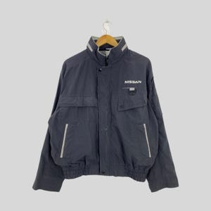 May include: A dark navy blue jacket with a high collar and multiple pockets. The jacket features the "NISSAN" logo in white on the chest. It has a zip closure and reflective silver stripes on the front and back.