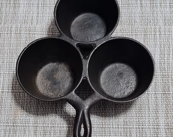 May include: A cast iron three-sectioned pan with a handle. Each section is round and has a slightly raised lip. The pan is black and has a rustic appearance.