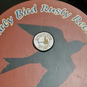 May include: A circular design with the text "Early Bird Rusty Relics" in blue lettering on a reddish-brown background. A black bird silhouette is visible. A circular patch in the center reads "Spring Lake Village Security."
