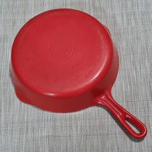 May include: Red cast iron skillet with a single loop handle. The skillet has a smooth, red enamel finish.