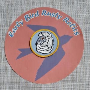 May include: A round, pink paper cutout with the text "Early Bird Rusty Relics" in blue script. A blue bird silhouette is visible. A circular patch with a sheep design is centered on the cutout. The patch has a blue background, a yellow border, and a white and gray sheep.