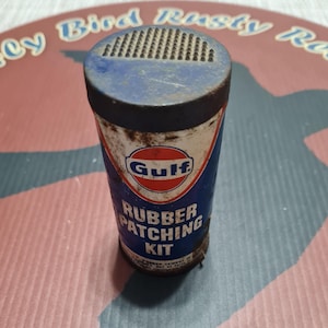 May include: Vintage Gulf Rubber Patching Kit tin. The cylindrical tin features a blue and white design with the Gulf logo in red. The text "Rubber Patching Kit" is displayed on the front. The tin has a perforated top and shows signs of age and wear.