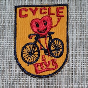 May include: A yellow and blue embroidered patch with a red heart riding a bicycle. The heart has a smiling face and the text "CYCLE & LIVE" is embroidered on the patch.