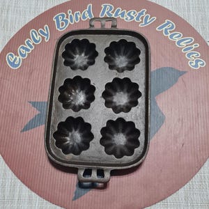 May include: A cast iron muffin pan with six flower-shaped molds. The pan has two handles and is set on a pink and red background with the text "Early Bird Rusty Relics".