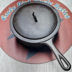 May include: A dark, seasoned cast iron skillet with a matching circular lid featuring a central handle. The pan has a long, integrated handle with a hole at the end for hanging. The surface of the metal is textured and matte black in appearance.
