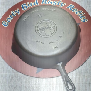 May include: A cast iron Griswold skillet, size 9, with a handle. The skillet is gray and sits on a red and brown circular background with the text "Early Bird Rusty Relics". The skillet is marked "Erie, PA., U.S.A."