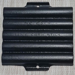 May include: Black cast iron corn stick pan with a rectangular shape and four rounded molds. The pan has two handles with holes for hanging. The text "NO 23-S" and "TINY CORN STICK" are embossed on the surface.