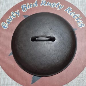 May include: A dark, cast iron pot lid with a loop handle. The lid is round and sits on a red and brown background with the text "Early Bird Rusty Relics" in blue script.