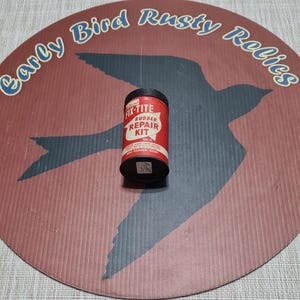 May include: A vintage Fix-Tite Rubber Repair Kit tin, red with white and black text, sits on a round, brown surface with a bird silhouette and the text "Early Bird Rusty Relics". The tin is cylindrical with a black lid.