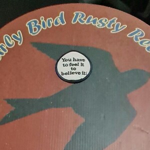 May include: A circular design with the text "Early Bird Rusty Relics" in white and blue lettering. A silhouette of a bird is visible, with a small circular sticker in the center that reads "You have to feel it to believe it!"