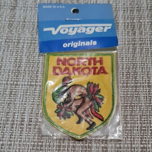 May include: A yellow embroidered patch with a green border. The patch features a cartoon image of a person in a green headdress, holding a red flower. The text "NORTH DAKOTA" is embroidered in red letters on the patch.